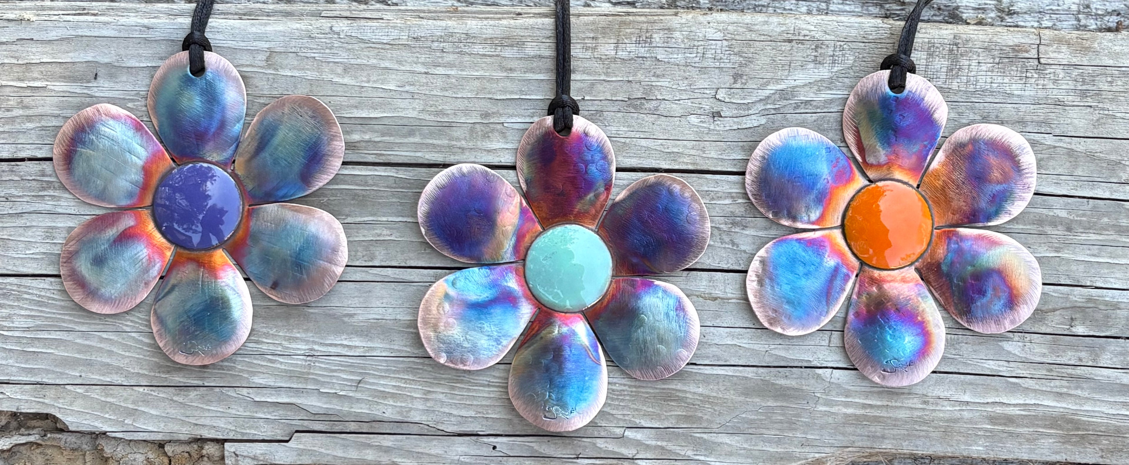 Three flower-shaped copper torch blown ornaments with multicolored glaze on a wooden surface.