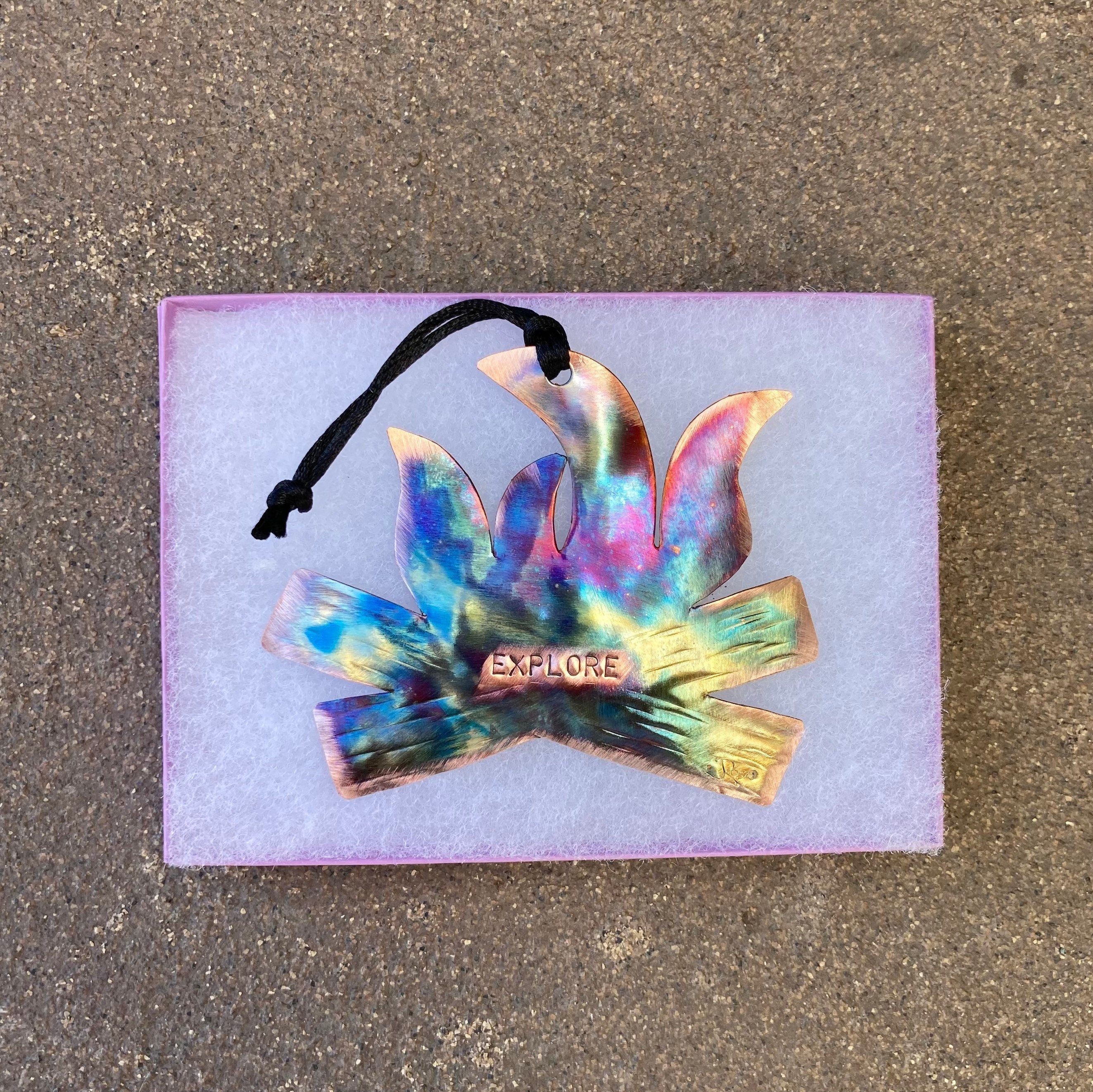 Colorful 'Explore' campfire design on a purple card with a black string, on a concrete surface.