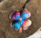 Colorful four-leaf clover pendant with a heart in the center on a textured surface.